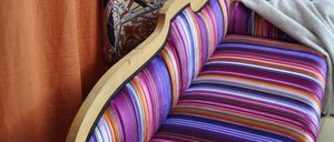 Nahaufnahme eines farbenfrohen gestreiften Sofas mit Holzrahmen. Das Sofa ist in leuchtenden Lila-, Rosa- und Orangetönen gepolstert. Eine weiße Decke liegt auf der Seite des Sofas.