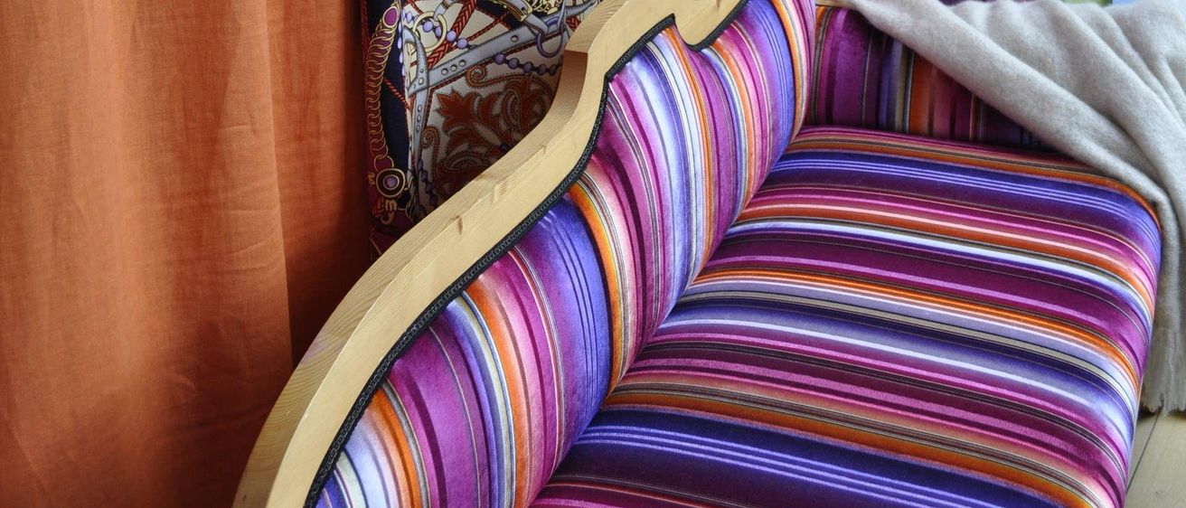 Nahaufnahme eines farbenfrohen gestreiften Sofas mit Holzrahmen. Das Sofa ist in leuchtenden Lila-, Rosa- und Orangetönen gepolstert. Eine weiße Decke liegt auf der Seite des Sofas.