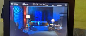 Ein Monitor zeigt ein Video eines Wohnzimmers mit blauem Licht. Das Videoformat ist 1080p25. Die Kameraeinstellungen zeigen ProRes HQ-Codec und SDI-Quelle. Der Bildschirm zeigt eine Zeit von 00:00:00:00.