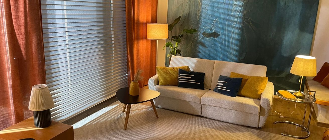 Innenansicht eines Wohnzimmers mit einem beigen Sofa und einem Tisch, einer Stehlampe und einem großen Gemälde an der Wand.