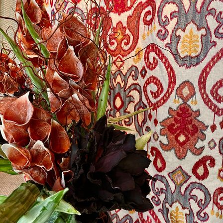 Ein Nahaufnahme einer Blumenarrangement mit roten und grünen Blättern und braunen Blumen vor einem rot-blau gemusterten Hintergrund.