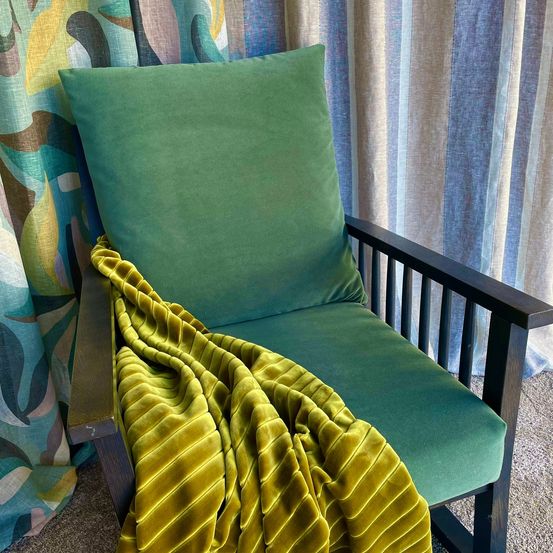 Ein grüner Samtstuhl mit einer gelben Samtdecke davor vor einem farbenfrohen und blau gestreiften Vorhang.