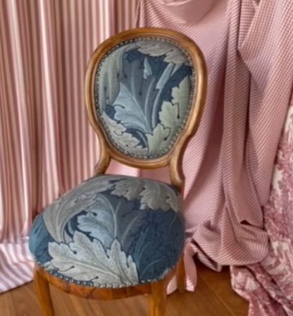 Ein Holzstuhl mit floralem Bezug steht vor einem rosa Vorhang mit Blumenmuster.