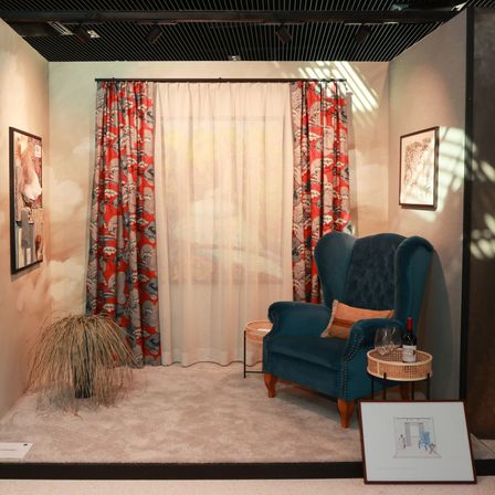 Ein Innenraum mit einem blauen Ohrensessel, einem weißen Vorhang, einem roten gemusterten Vorhang und gerahmten Bildern an der Wand.
