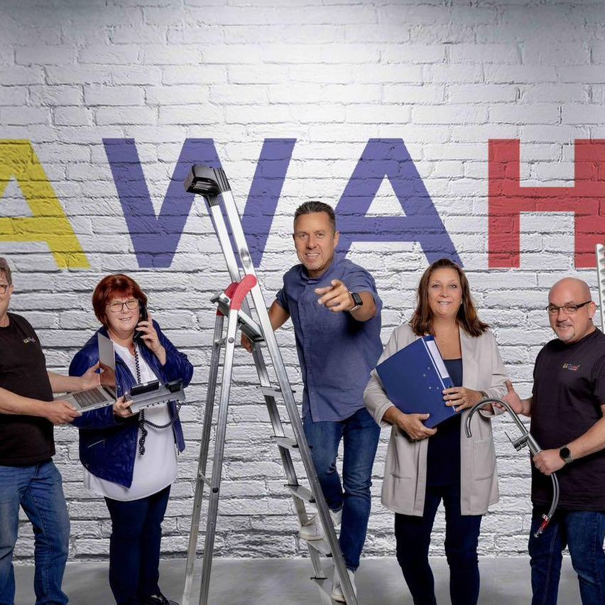 Eine Gruppe von Menschen steht vor einer Backsteinwand mit dem Wort "AWAHE". Ein Mann steht auf einer Leiter und hält ein Gerät. Die anderen haben verschiedene Werkzeuge und Dokumente.