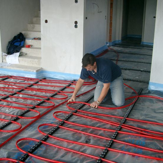 Ein Mann arbeitet auf dem Boden und installiert rote Rohre auf einer grauen Plastikfolie in einem unfertigen Raum.