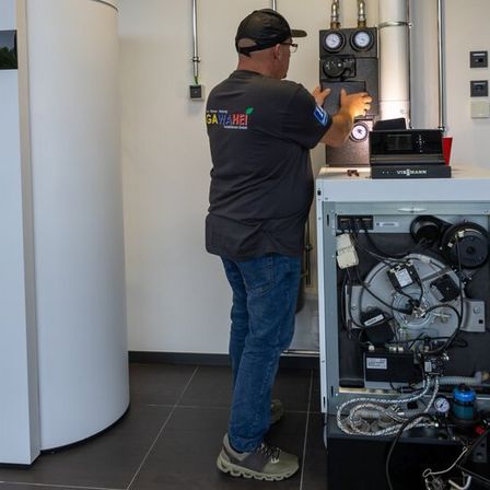 Ein Mann in einem schwarzen Shirt und Jeans justiert eine komplexe Industriemaschine in einem weißen Raum. In der Nähe befinden sich ein großer weißer Zylinder und ein Feuerlöscher.