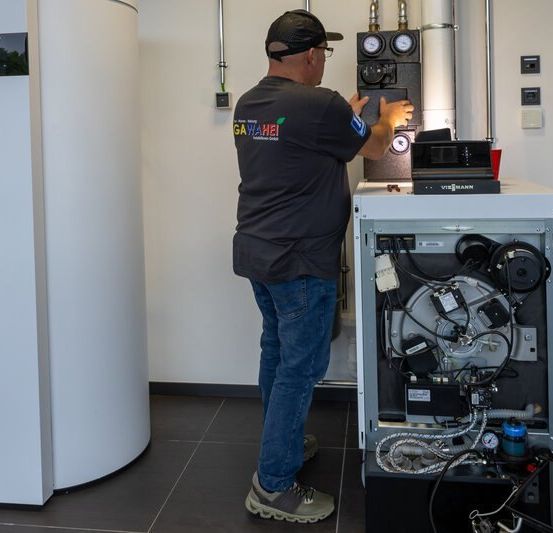 Ein Mann in einem schwarzen Shirt und Jeans justiert eine komplexe Industriemaschine in einem weißen Raum. In der Nähe befinden sich ein großer weißer Zylinder und ein Feuerlöscher.