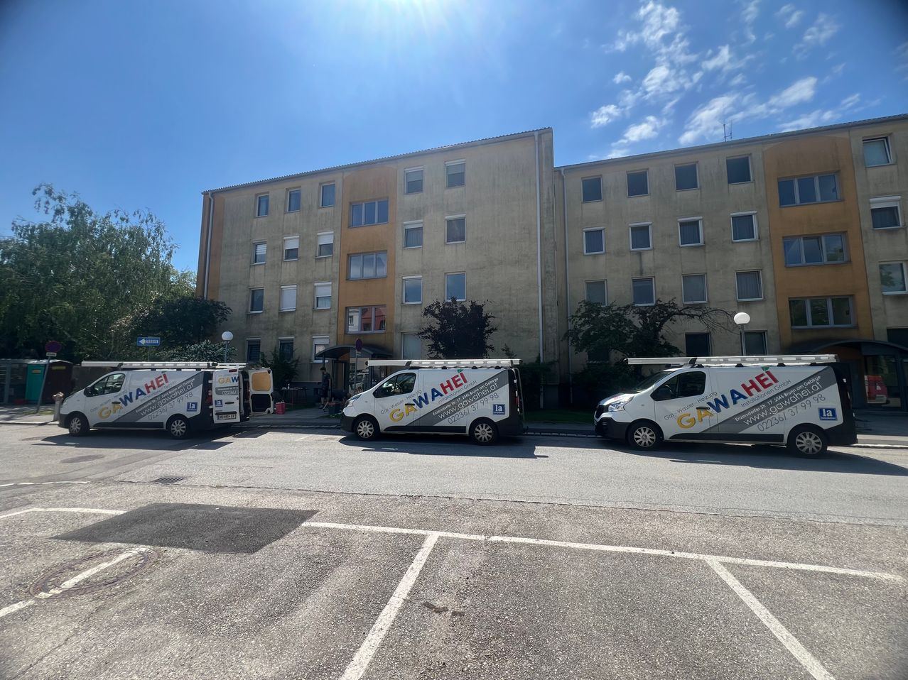 Three white vans with Gawahi on the side are parked in a parking lot in front of a large building. The building has many windows and a yellow wall.