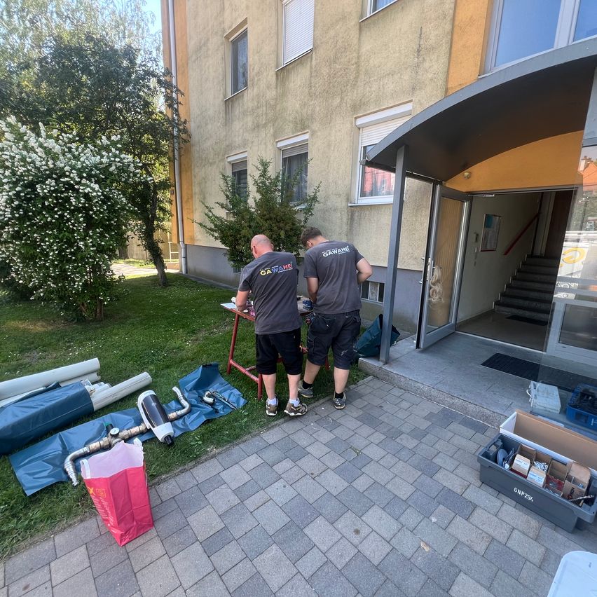 Two men in gray shirts work outside a building, near a table with tools and a red bag. Nearby is a grassy area with a tree and bushes.