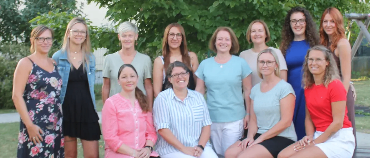 Eine Gruppe von Frauen posiert für ein Foto im Freien. Einige sitzen auf einer Bank, während andere stehen. Sie alle lächeln und schauen in die Kamera.