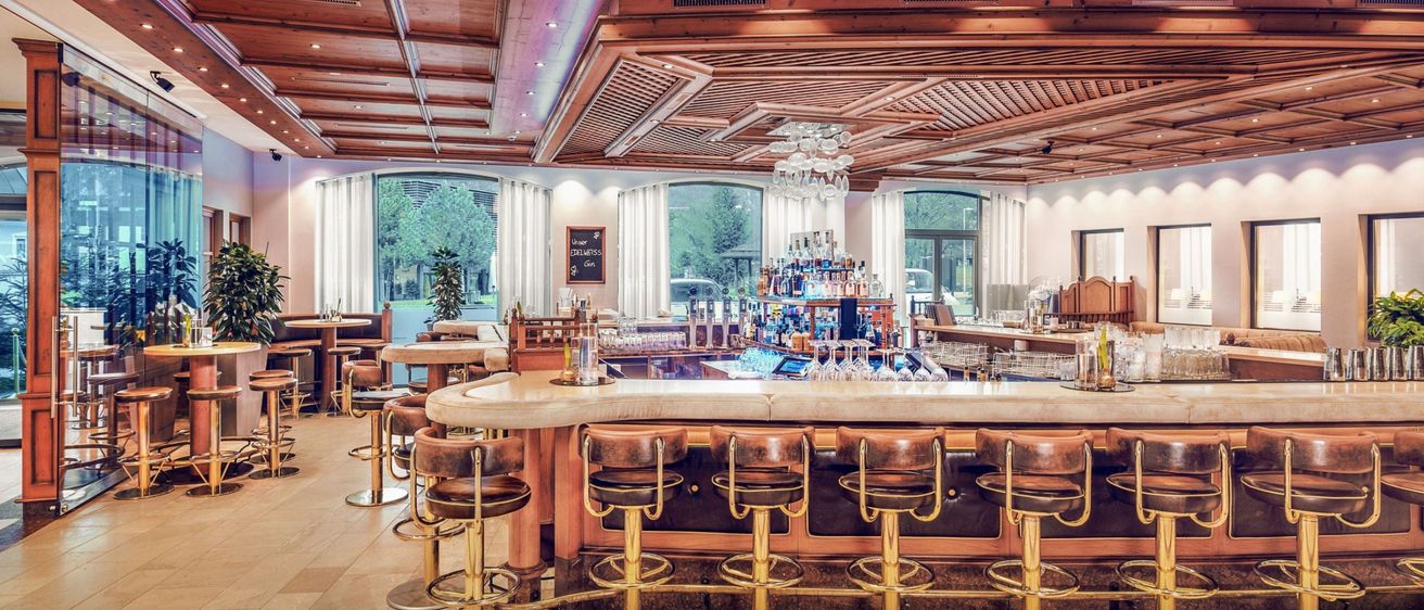 Ein moderner Barbereich mit Holzdecke und Glasfenstern, ausgestattet mit einer großen Theke, Barhockern, einer Kristallleuchte und Regalen mit Flaschen und Gläsern.