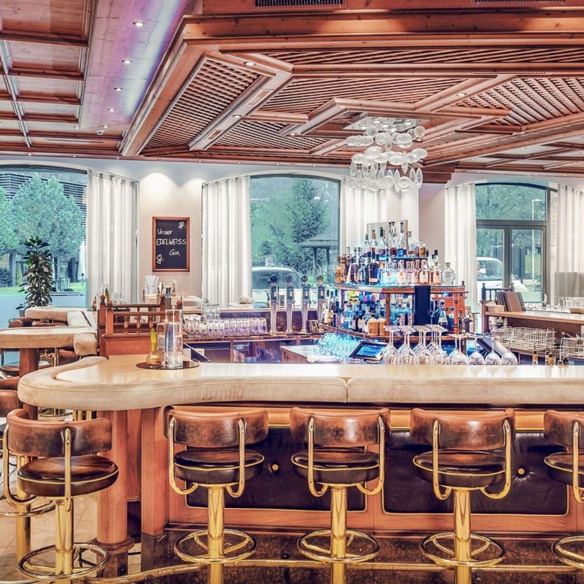 Ein moderner Barbereich mit Holzdecke und Glasfenstern, ausgestattet mit einer großen Theke, Barhockern, einer Kristallleuchte und Regalen mit Flaschen und Gläsern.