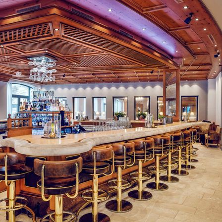 Eine luxuriöse Bar mit einer geschwungenen Marmortheke, Lederstühlen und einer Holzdecke. Die Bar hat verschiedene Flaschen, Gläser und Pflanzen. Es hängen zwei Kronleuchter an der Decke. Daneben steht eine Couch mit einem Kissen und einer Lampe.