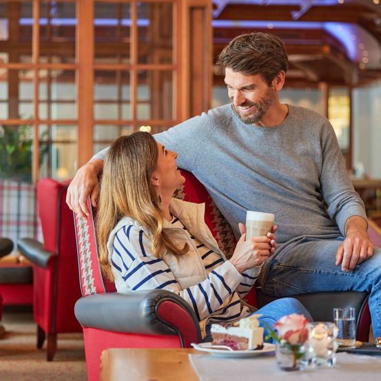 Ein Paar genießt einen gemütlichen Moment in einem Café, sitzt auf einem roten Sessel, wobei der Mann eine Tasse hält und die Frau ihn ansieht. Sie sind umgeben von einem Tisch mit Kuchen, einer Kerze und einem Glas Wasser. Hinter ihnen sind Holzwände und ein roter Stuhl.