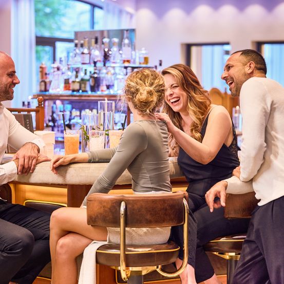 Eine Gruppe von Menschen sitzt an einer Bar, lächelt und lacht. Auf der Theke stehen Gläser mit Getränken. Hinter ihnen ordnet ein Barkeeper Flaschen.