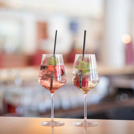 Zwei Weingläser mit Früchten und Minzgarnierung auf einem Holztisch mit unscharfem Hintergrund.