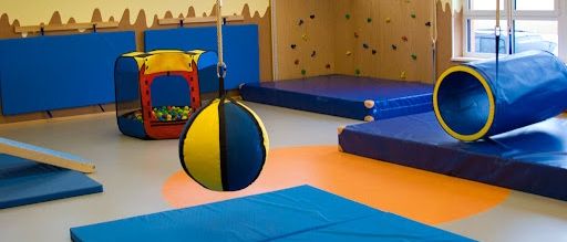 Ein Spielzimmer mit blauen und gelben Schaumstoffmatten, einer Schaukel und einer Kletterwand mit verschiedenfarbigen Griffen.