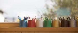 Fünf kleine Stifthalter in verschiedenen Farben sind auf einem Holzregal neben einem Fenster angeordnet. Jeder Halter enthält eine Gruppe von Stiften.
