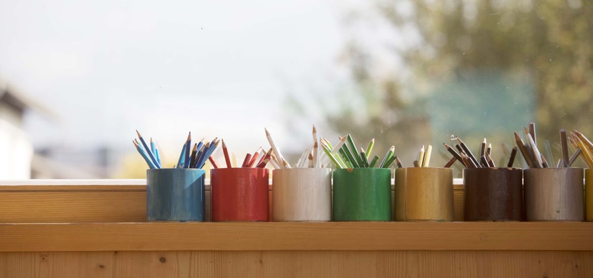 Fünf kleine Stifthalter in verschiedenen Farben sind auf einem Holzregal neben einem Fenster angeordnet. Jeder Halter enthält eine Gruppe von Stiften.