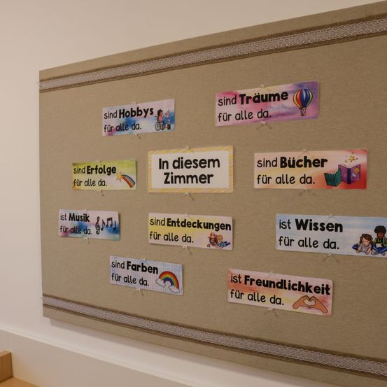 Ein Bulletinbrett mit mehreren bunten Schildern. Jedes Schild hat ein anderes Thema wie Hobbys, Bücher und Entdeckungen. Alle Schilder haben die Phrase 'für alle da' auf Deutsch.