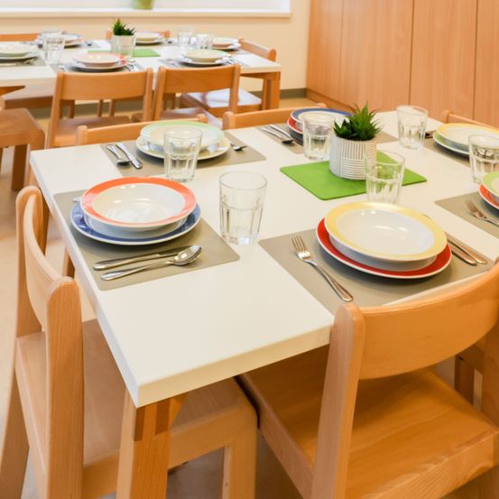 Ein Esszimmer mit mehreren hölzernen Stühlen um einen großen weißen Tisch. Jede Platz-Einrichtung umfasst bunte Teller, Gläser und Besteck. Ein Topfpflanzen und ein kleines grünes Tischset sind auf dem Tisch.