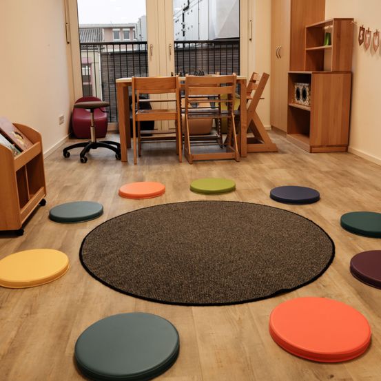 Ein Raum mit Holzböden, einem runden Teppich und gepolsterten Sitzen darum herum. Ein Esstisch mit Stühlen ist aufgestellt, und ein Bücherregal steht an der Wand. Ein Balkon mit Geländer ist durch das Fenster sichtbar.