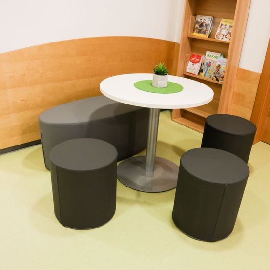 Eine kleine Sitzecke mit einem runden Tisch und vier schwarzen Hockern, einem Topf mit Pflanzen auf dem Tisch und einem hölzernen Bücherregal dahinter.