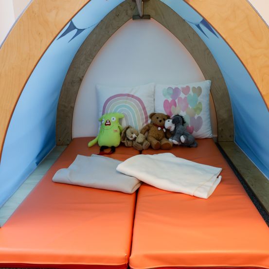 Ein orangefarbenes Bett in einem Zelt mit zwei Kissen, eines mit Regenbogendesign und eines mit Herzdesigns. Gefüllte Plüschtiere liegen auf dem Bett, zusammen mit zwei Decken.