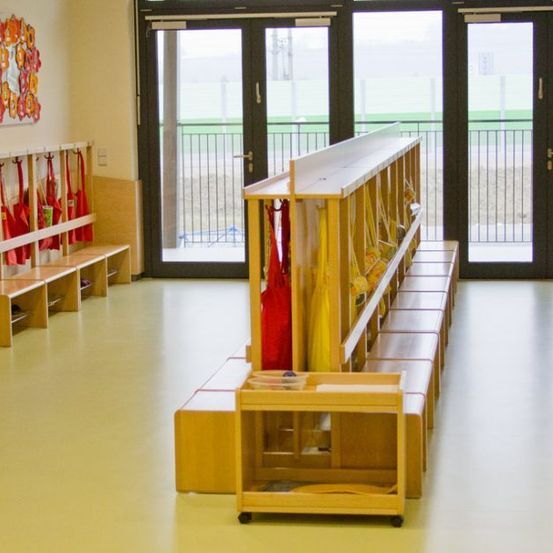 Ein leerer Raum mit Holzbänken und Regalen, an denen rote Taschen hängen. Eine bunte Wanddekoration befindet sich an der linken Wand. Der Raum hat große Glastüren, die zu einem Außenbereich führen.