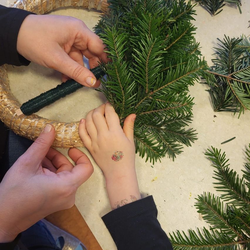 Zwei Hände arbeiten an einem Kranz, eine davon hat ein Blumentattoo am Handgelenk. Der Kranz besteht aus grünen Tannennadeln.