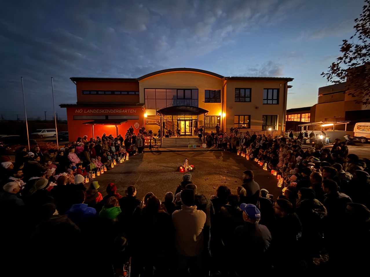 Eine Menschenmenge versammelt sich nachts um einen Kindergarten und hält kreisförmig brennende Kerzen. Das Gebäude hat große Fenster und ein Schild, das NO LANDESKINDGARTEN steht.