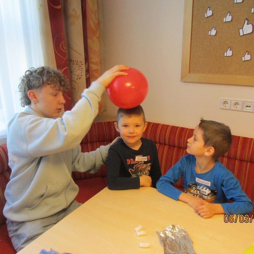 Ein Junge balanciert einen roten Ballon auf dem Kopf eines anderen Jungen, während sie um einen Tisch mit zwei anderen Jungen sitzen. Eine Pinnwand mit Daumen-hoch-Bildern ist an der Wand montiert.