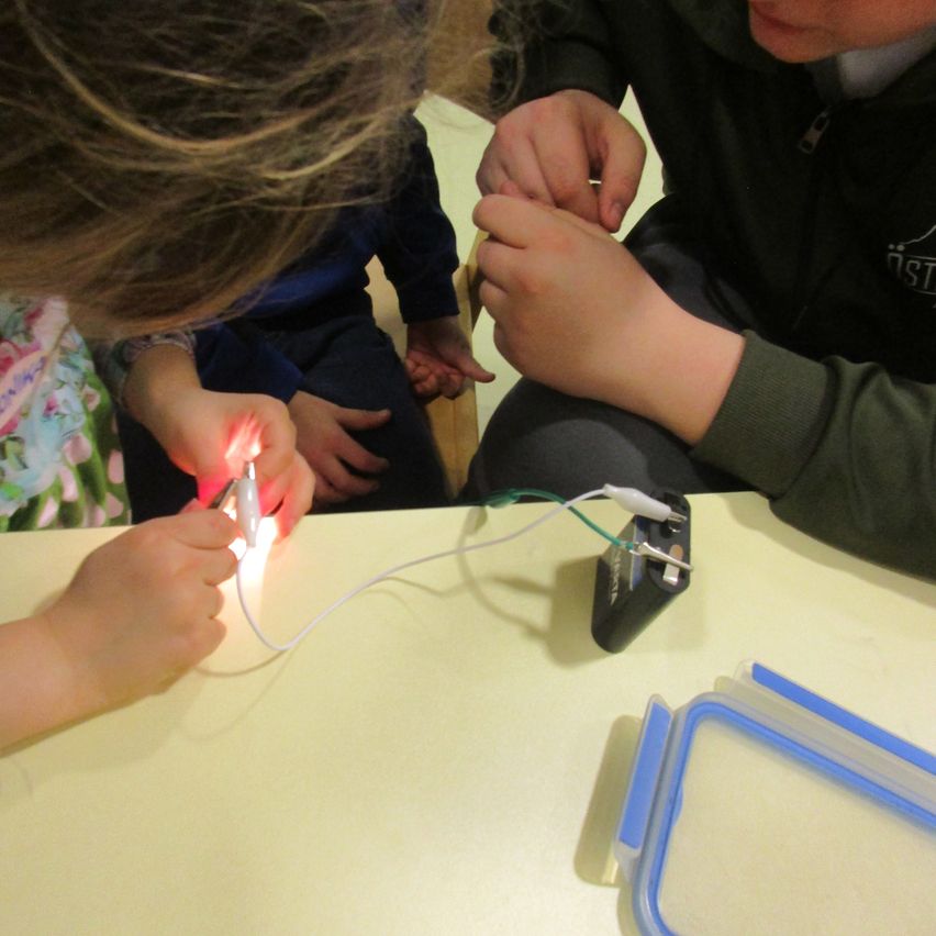 Eine Gruppe von Kindern experimentiert mit einer leuchtenden Taschenlampe, die an eine Batterie angeschlossen ist. Ein Kind hält die Taschenlampe und andere beobachten aufmerksam.