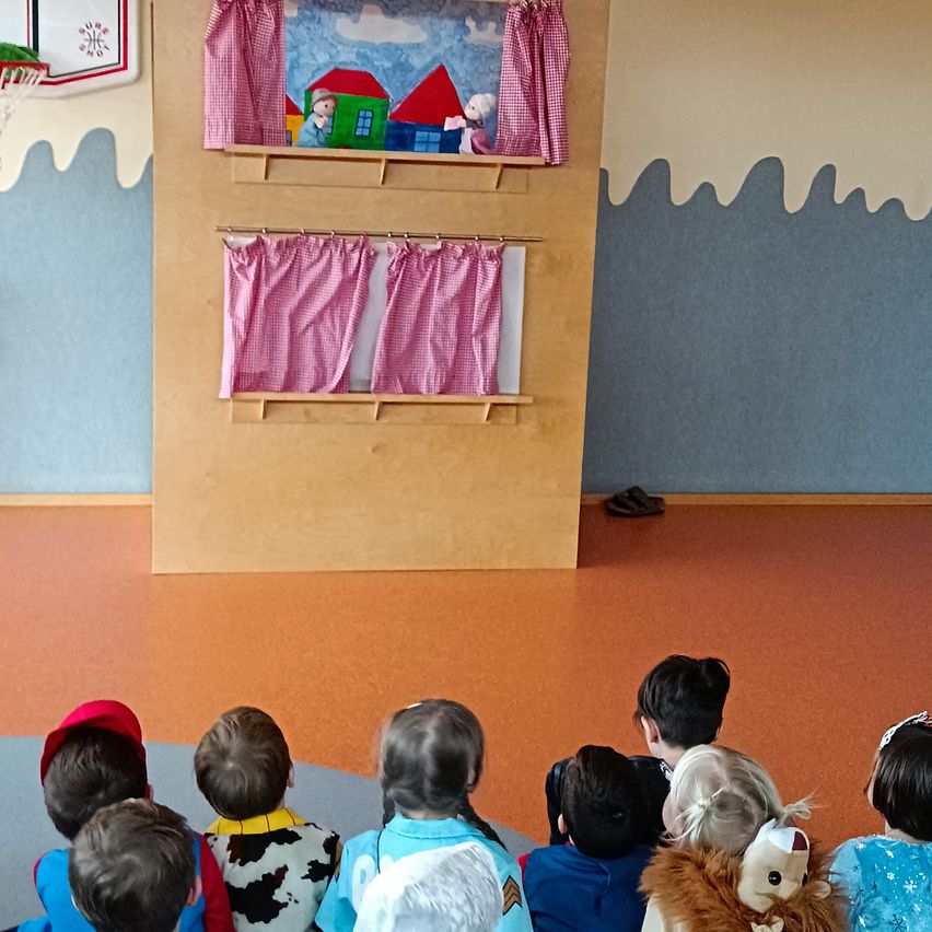 Eine Gruppe von Kindern sieht sich ein Puppenspiel mit einem Puppentheater im Klassenzimmer an. Im Hintergrund befinden sich ein roter Vorhang und eine Wand mit einem Basketballkorb.