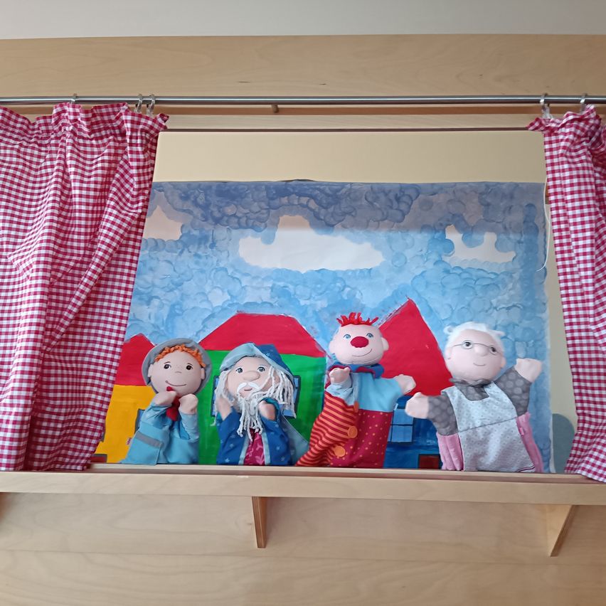 Vier Puppen stehen auf einer Bühne mit einem Hintergrund, der mit Wolken und Häusern bemalt ist. Die Puppen sind in verschiedenen Kostümen gekleidet. Rote und weiße Vorhänge hängen an einer Metallstange über der Bühne.