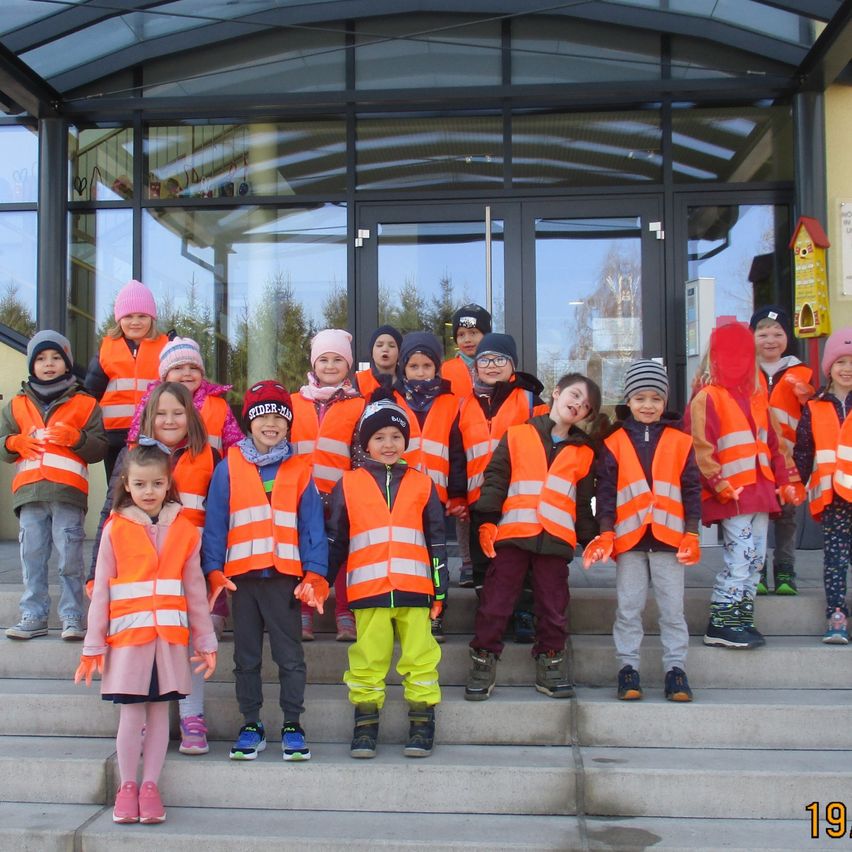 Eine Gruppe junger Kinder in Warnwesten steht auf Stufen vor einem Gebäude. Im Hintergrund sind Bäume zu sehen.
