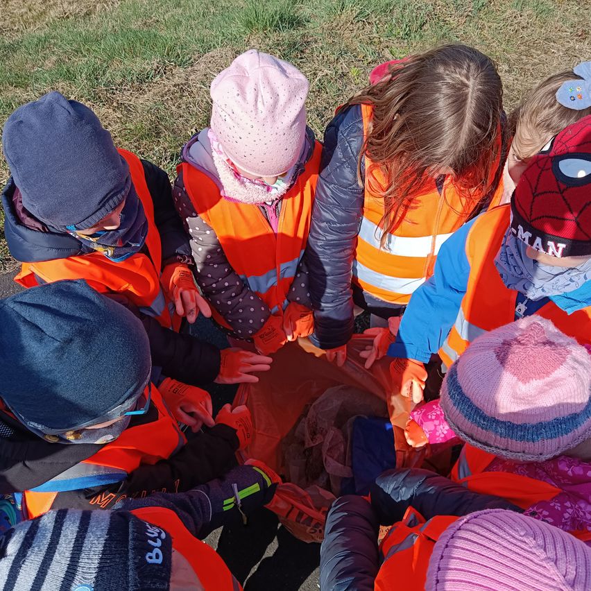 Kinder in Warnwesten und Mützen sammeln sich um einen Beutel, möglicherweise Müll aufsammelnd. Der Boden ist mit trockenem Gras bedeckt.
