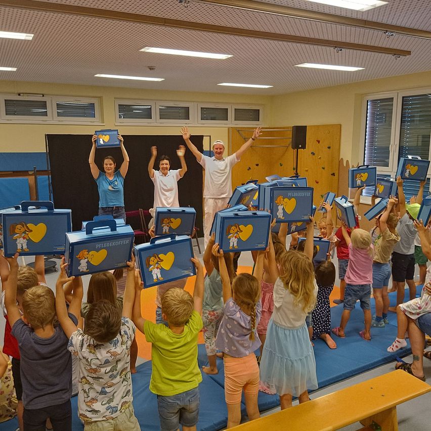 Eine Gruppe von Kindern in einem Raum mit blauem Teppich halten Kisten hoch. Zwei Erwachsene stehen vorne mit erhobenen Händen.