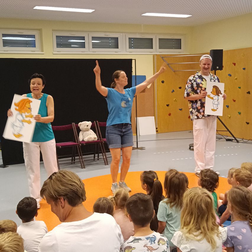 Drei Personen stehen auf der Bühne und halten Zeichnungen in die Höhe. Eine hebt die Arme. Dahinter befindet sich eine Kletterwand und ein schwarzer Hintergrund. Kinder sitzen vor ihnen auf dem Boden.