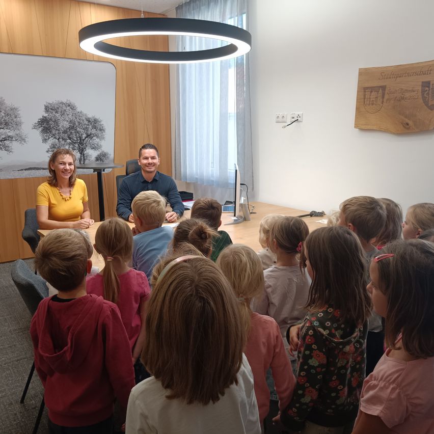 Eine Gruppe von Kindern ist um einen Tisch versammelt, an dem zwei Erwachsene sitzen. Der Raum hat Holzwände und ein großes Fenster mit weißen Vorhängen.