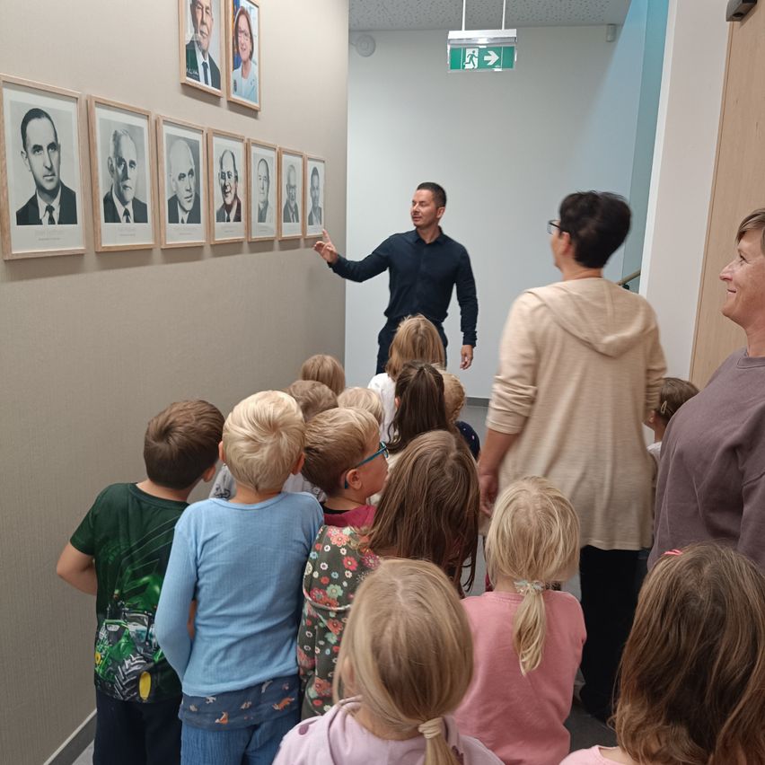 Ein Mann hält eine Präsentation für eine Gruppe von Kindern in einem Flur mit Porträts historischer Persönlichkeiten an der Wand.
