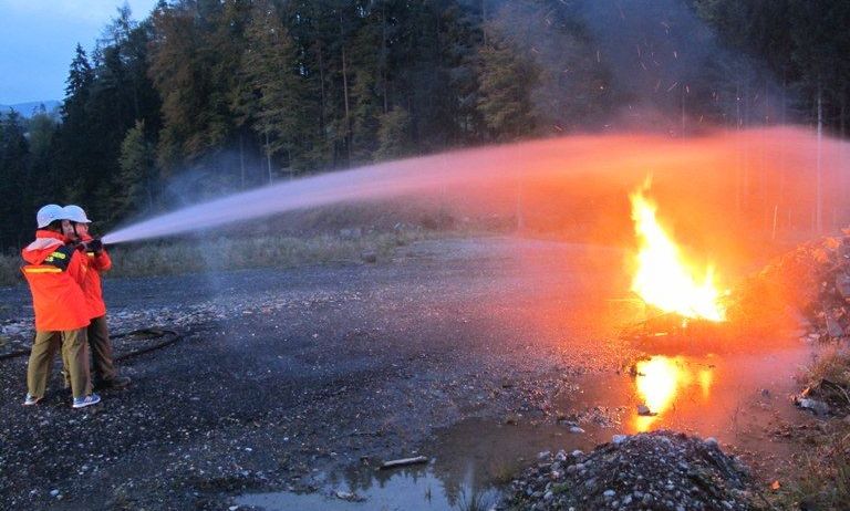 Zwei Feuerwehrleute verwenden einen Schlauch, um ein Feuer in einem Außenbereich mit einem Schotterweg zu löschen.