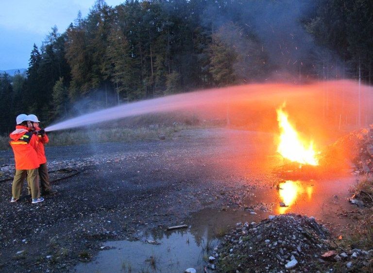 Zwei Feuerwehrleute verwenden einen Schlauch, um ein Feuer in einem Außenbereich mit einem Schotterweg zu löschen.