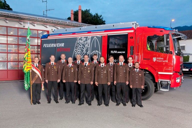 Eine Gruppe von Feuerwehrleuten posiert vor einem Feuerwehrauto mit den Aufschriften 'Feuerwehr' und 'TLFA 3000/100'. Sie tragen Uniformen und Mützen.