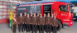 Eine Gruppe von Feuerwehrleuten posiert vor einem Feuerwehrauto mit den Aufschriften 'Feuerwehr' und 'TLFA 3000/100'. Sie tragen Uniformen und Mützen.