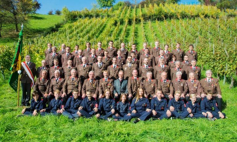 Eine Gruppe uniformierter Personen, einige stehend und einige sitzend, posiert für ein Foto in einem Weinberg unter klarem Himmel.