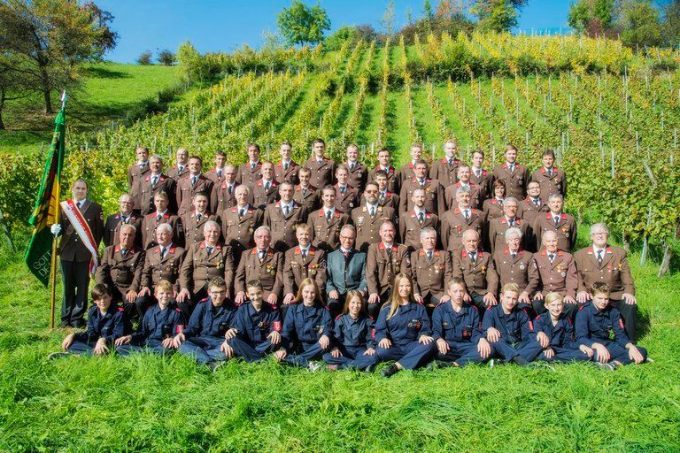 Eine Gruppe uniformierter Personen, einige stehend und einige sitzend, posiert für ein Foto in einem Weinberg unter klarem Himmel.