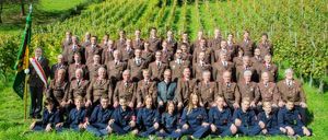 Eine Gruppe uniformierter Personen, einige stehend und einige sitzend, posiert für ein Foto in einem Weinberg unter klarem Himmel.