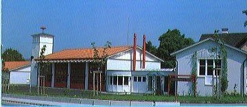 Ein Bild eines modernen Gebäudes mit einem roten Dach, umgeben von Grün und einem Schwimmbad davor. Es gibt zwei hohe Stangen an der Seite. Bäume und ein weiteres Gebäude sind im Hintergrund zu sehen.